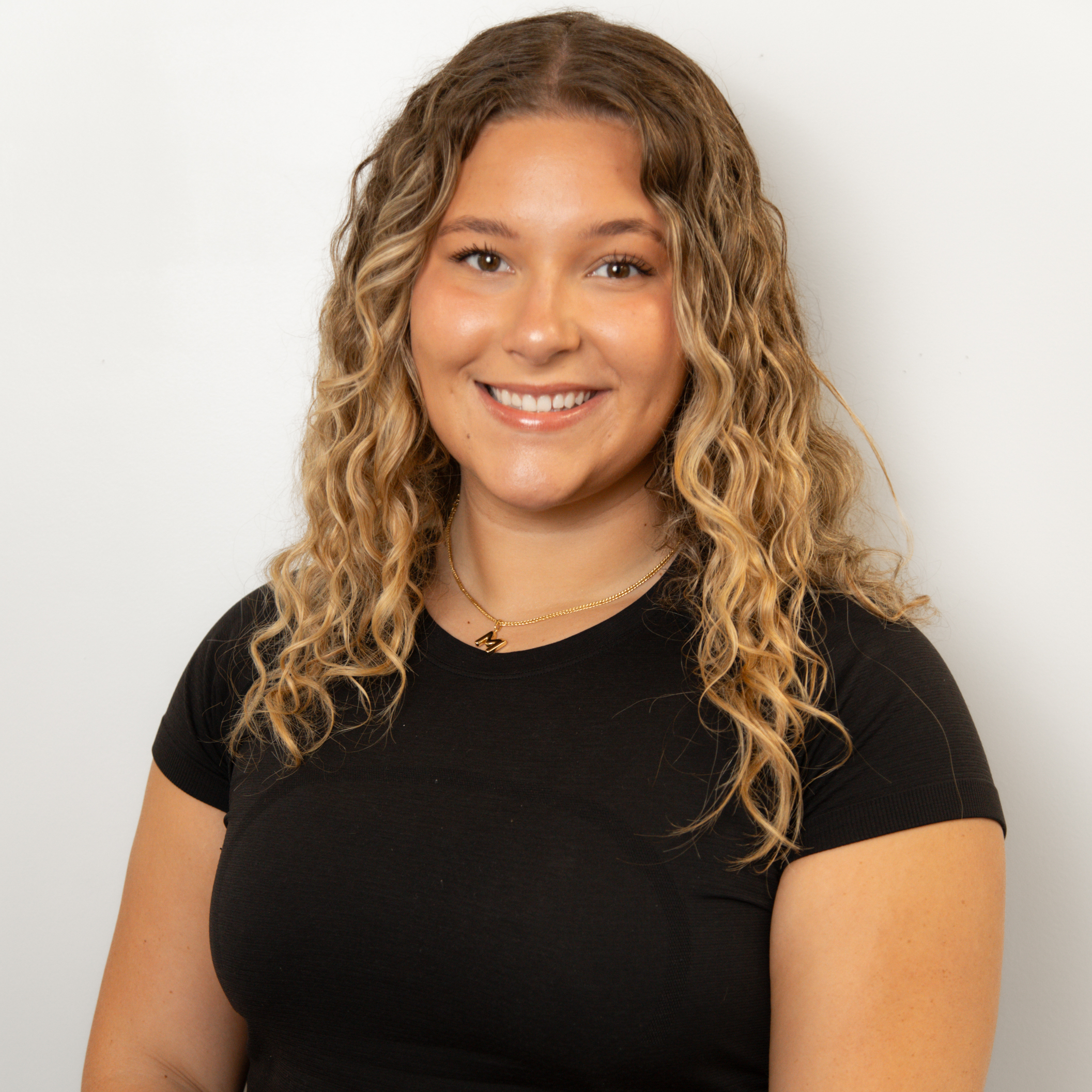 Smiling woman with long, curly blonde hair wearing a black top, posed against a light wall.
